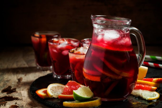 A imagem mostra uma jarra com sangria e alguns pedaços de frutas e gelo, além de copos de vidro, sobre uma mesa.