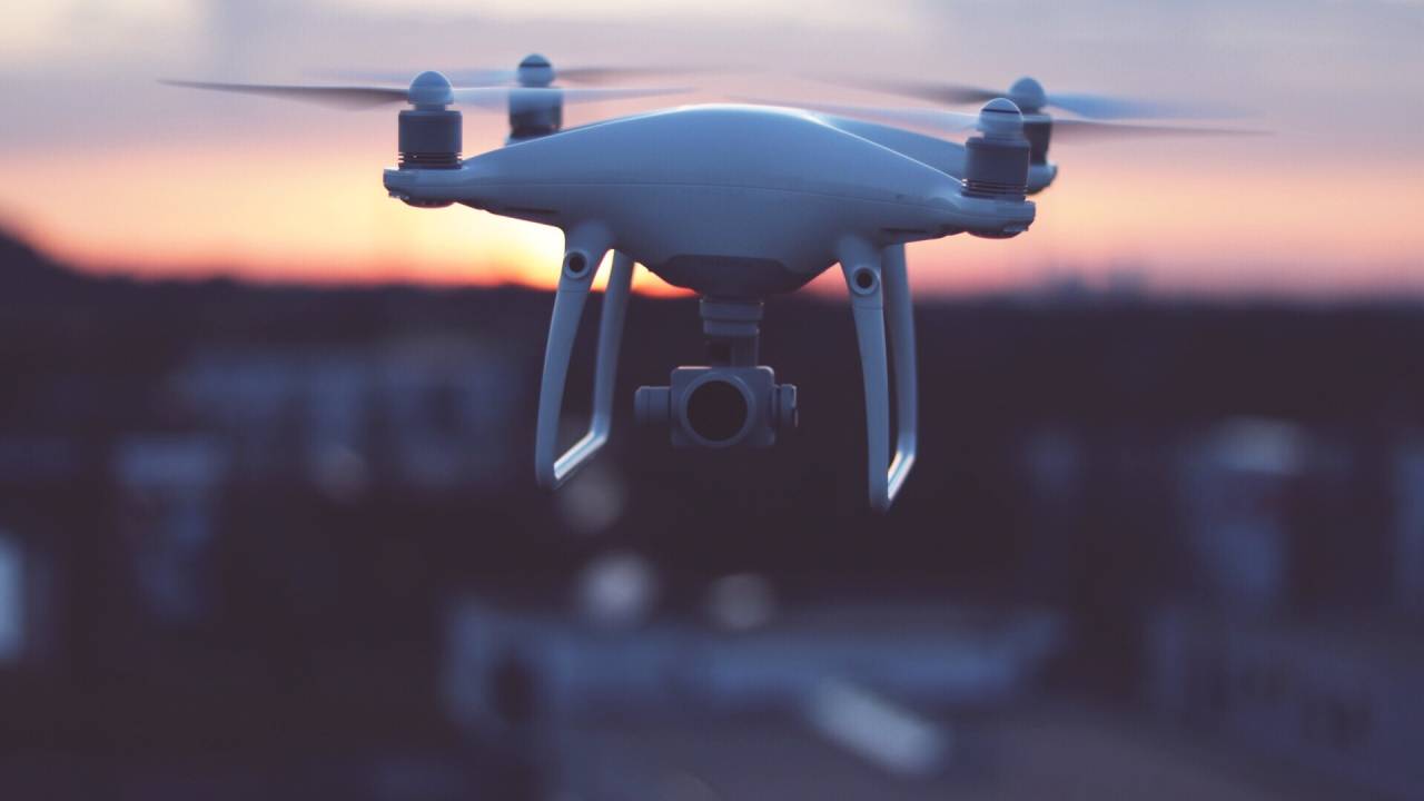 Drone na cor branca voando. Ao fundo é possível ver o céu com o sol se pondo em tons de laranja.