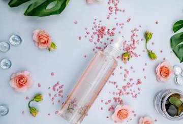 Frasco de perfume de cor rosada, num fundo azul claro, cercado por flores e folhas.