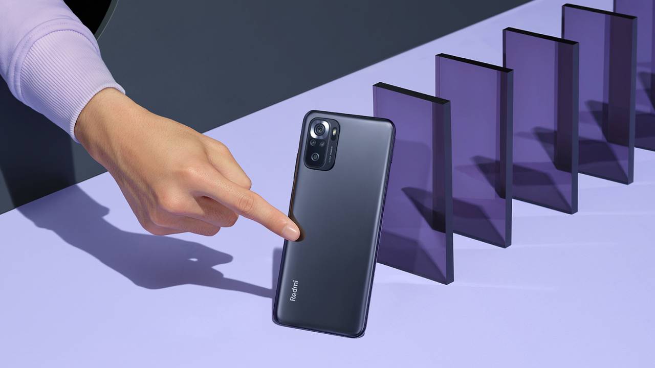 A imagem mostra o Redmi Note 10S, num fundo lilás, com um dedo empurrando o smartphone e, atrás dele, diversas peças de acrílico roxas, do mesmo tamanho do celular, em pé, alinhadas formando uma fila.