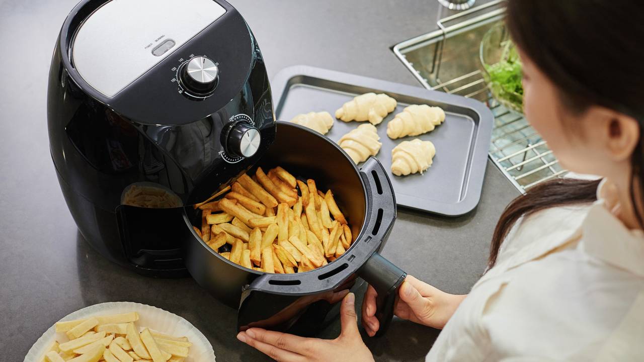 A imagem mostra uma mulher de costas, abrindo uma air fryer cheia de batatas fritas, em cima de um balcão.