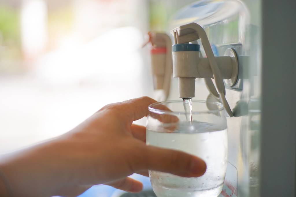 A imagem mostra uma mão, pegando água de um purificador de água com um copo de vidro.