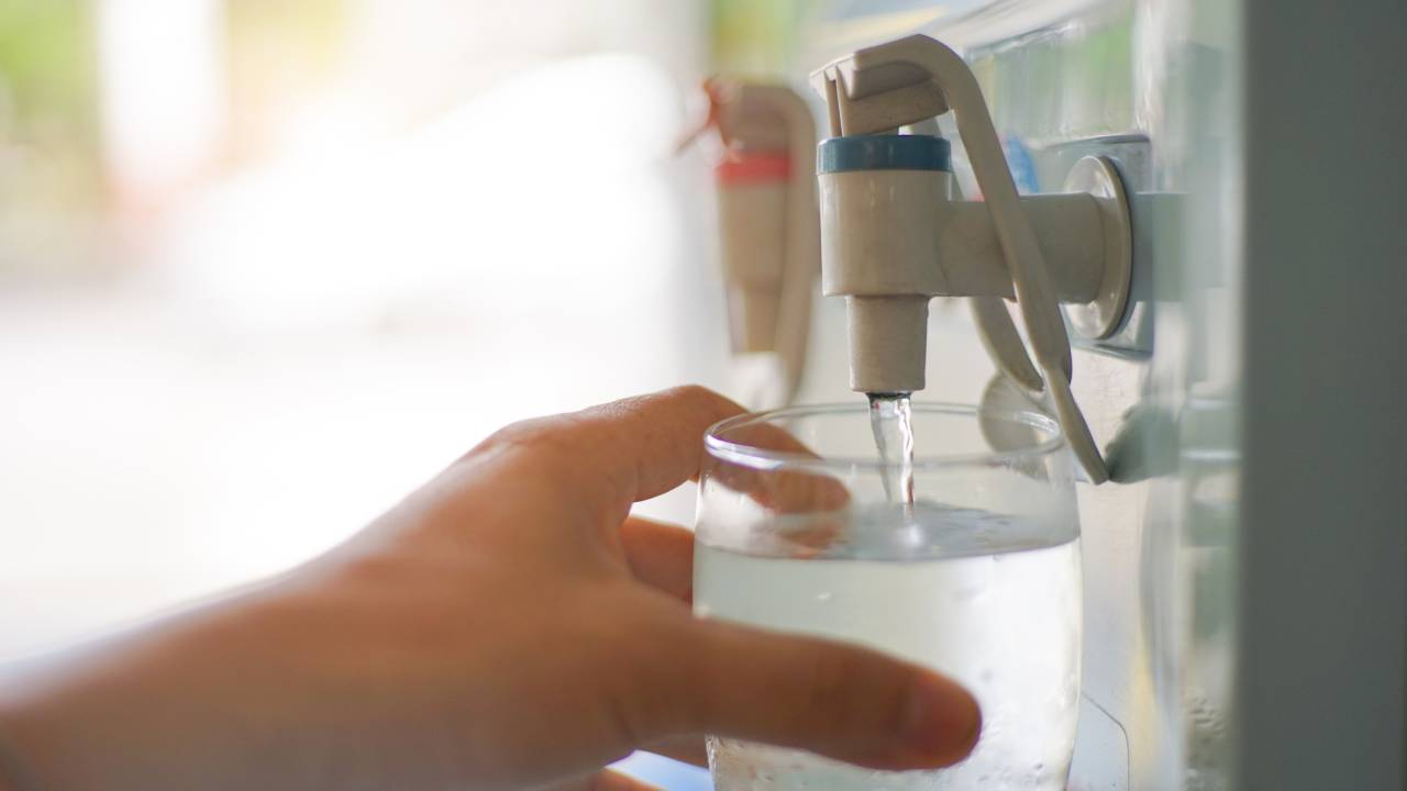A imagem mostra uma mão, pegando água de um purificador de água com um copo de vidro.