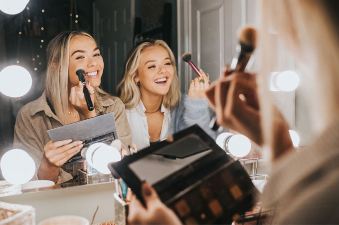 Duas mulheres se maquianda em frente ao espelho e rindo