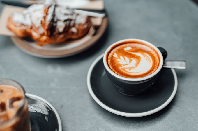 Café espresso com espuma de leite sobre a mesa, onde se vê também um croissant e um chocolate quente