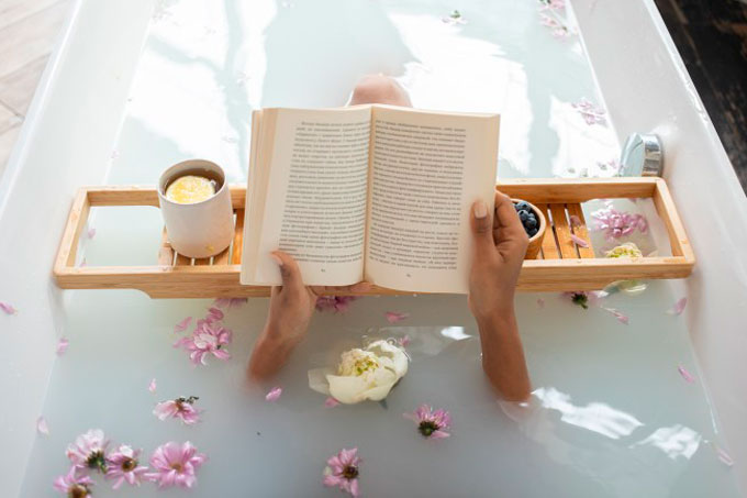 Dentro de uma banheira com água e flores, vemos duas mãos femininas lendo um livro, com apoio numa bandeja de madeira