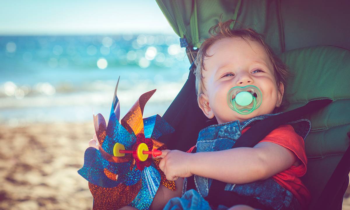 bebê sorri com chupeta na boca, dentro de um carrinho na praia